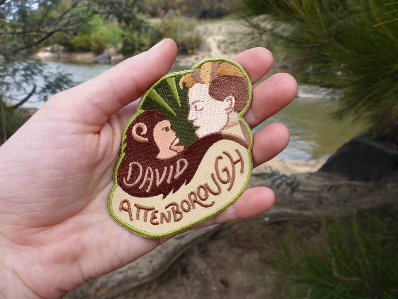 K&ouml;nnte beinhalten: Ein gestickter Aufn&auml;her in Gr&uuml;n, Braun und Beige mit einem Cartoon-Bild eines Mannes mit einem Affen. Der Text "David Attenborough" ist auf den Aufn&auml;her gestickt.