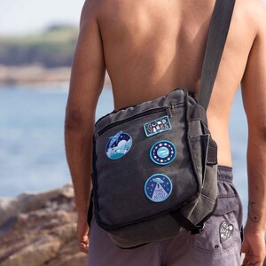 May include: A person wearing grey shorts with a black canvas messenger bag with various patches. The patches include a blue and white planet, a blue and white mountain scene, a blue and white UFO, and a blue and white patch with the text "Take The Rest Now".