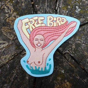 Puede incluir: Parche bordado con una mujer de pelo rosa buceando en agua azul con el texto "FREE BIRD" en amarillo.