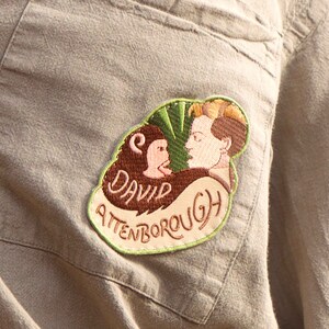 K&ouml;nnte beinhalten: Ein beigefarbener Stoff-Patch mit gr&uuml;nen, braunen und rosafarbenen Stickereien. Der Patch zeigt eine Karikatur einer Person mit einem Affen und den Text "David Attenborough".