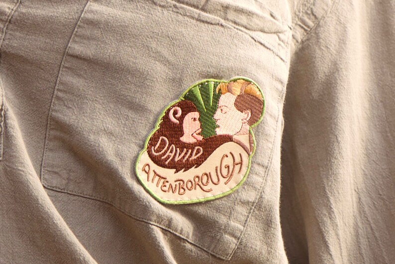 K&ouml;nnte beinhalten: Ein beigefarbener Stoff-Patch mit gr&uuml;nen, braunen und rosafarbenen Stickereien. Der Patch zeigt eine Karikatur einer Person mit einem Affen und den Text "David Attenborough".