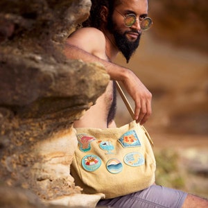 May include: A tan canvas tote bag with a variety of colorful embroidered patches. The patches feature images of a seagull, a cocktail, a wave, a crab, and a fish. The bag is worn by a person with a beard and sunglasses.