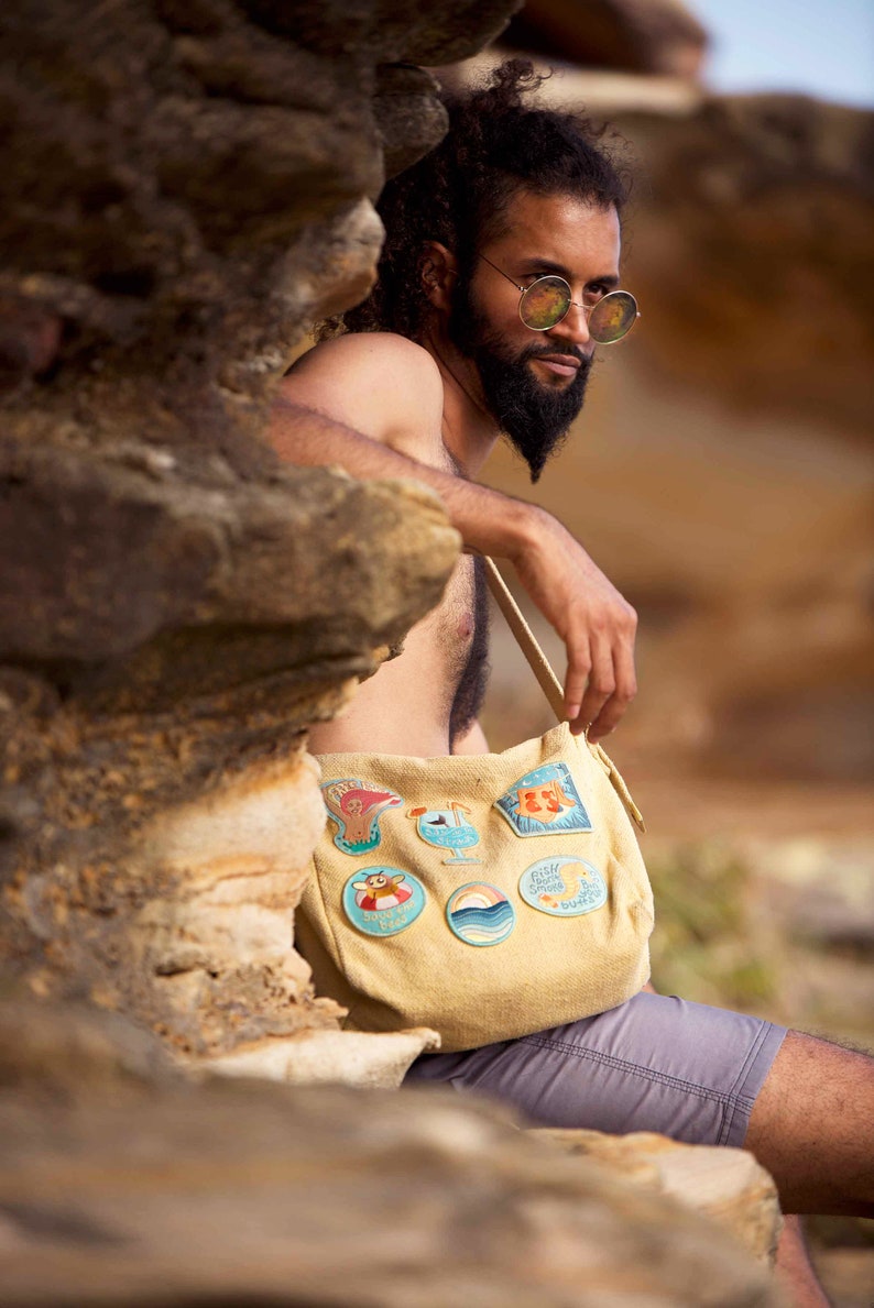 May include: A tan canvas tote bag with a variety of colorful embroidered patches. The patches feature images of a seagull, a cocktail, a wave, a crab, and a fish. The bag is worn by a person with a beard and sunglasses.
