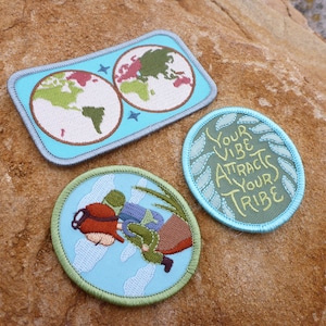 Può includere: Tre toppe ricamate con sfondo blu e cuciture verdi. Una toppa presenta una mappa del mondo con continenti rossi, verdi e marroni. Un'altra toppa presenta un'illustrazione di cartoni animati di una persona con uno zaino e occhiali protettivi che vola nell'aria. La terza toppa ha il testo "Your vibe attracts your tribe" in lettere gialle.