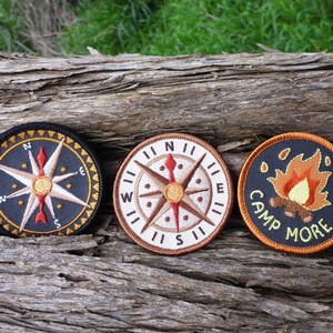 May include: Three embroidered patches on a wooden surface. The patches are circular and feature a compass, a compass with a red arrow, and a campfire with the text "Camp More".