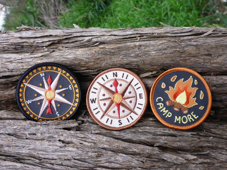 May include: Three embroidered patches on a wooden surface. The patches are circular and feature a compass, a compass with a red arrow, and a campfire with the text "Camp More".
