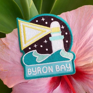 May include: A teal blue embroidered patch featuring a lighthouse, waves, and a yellow triangle. The patch is labelled "BYRON BAY" in white.