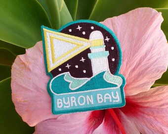 Op de afbeelding: Een geborduurde patch in turquoise blauw met een vuurtoren, golven en een gele driehoek. De patch heeft de tekst "BYRON BAY" in wit.