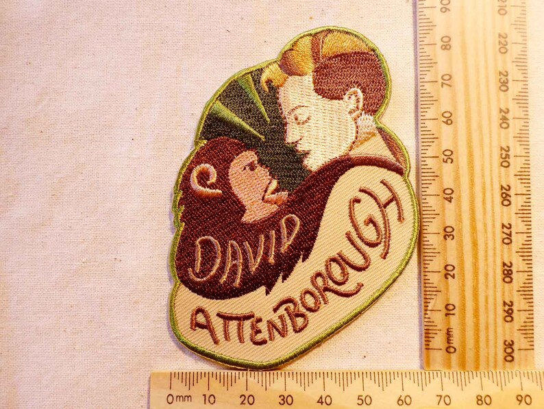 K&ouml;nnte beinhalten: Ein gestickter Aufn&auml;her mit einem Cartoon-Bild eines Mannes und eines Affen mit dem Text "David Attenborough" in brauner, wei&szlig;er und gr&uuml;ner Schrift.