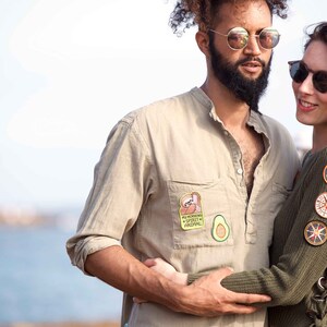 May include: A couple wearing casual clothing with patches on their shirts. The man is wearing a beige shirt with a sloth patch and an avocado patch. The woman is wearing a green sweater with several patches, including a compass and a campfire.