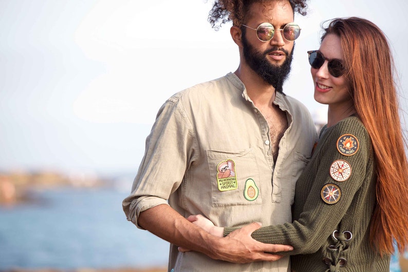 May include: A couple wearing casual clothing with patches on their shirts. The man is wearing a beige shirt with a sloth patch and an avocado patch. The woman is wearing a green sweater with several patches, including a compass and a campfire.