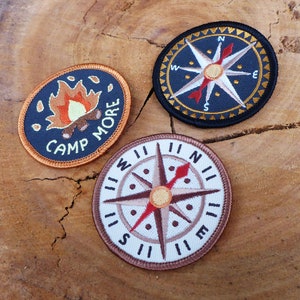 May include: Three embroidered patches with a camping theme. One patch is black with a compass design and the words "N", "W", "E", and "S". Another patch is black with a campfire design and the words "Camp More". The third patch is white with a compass design and the words "N", "W", "E", and "S" in Roman numerals.