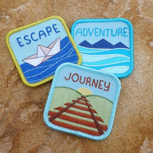 May include: Three embroidered iron-on patches with blue backgrounds and white stitching. The patches feature the words "Escape", "Adventure", and "Journey" with corresponding images. The "Escape" patch has a paper boat on blue water. The "Adventure" patch has blue mountains and waves. The "Journey" patch has a brown railway track with a green field and a yellow sun.