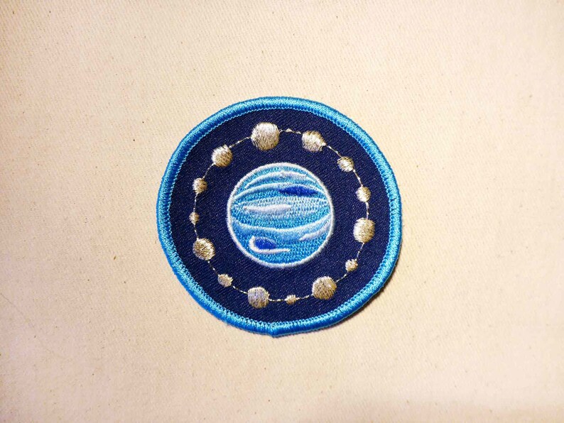 May include: An embroidered patch featuring a blue and white planet design, encircled by a ring of smaller circles. The patch is displayed against a beige fabric backdrop.
