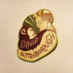 K&ouml;nnte beinhalten: Ein gestickter Aufn&auml;her in Gr&uuml;n, Braun und Beige mit einer Cartoon-Abbildung eines Mannes mit einem Affen auf der Schulter. Der Text "David Attenborough" ist in einer kursiv geschriebenen Schrift auf dem Aufn&auml;her gestickt.
