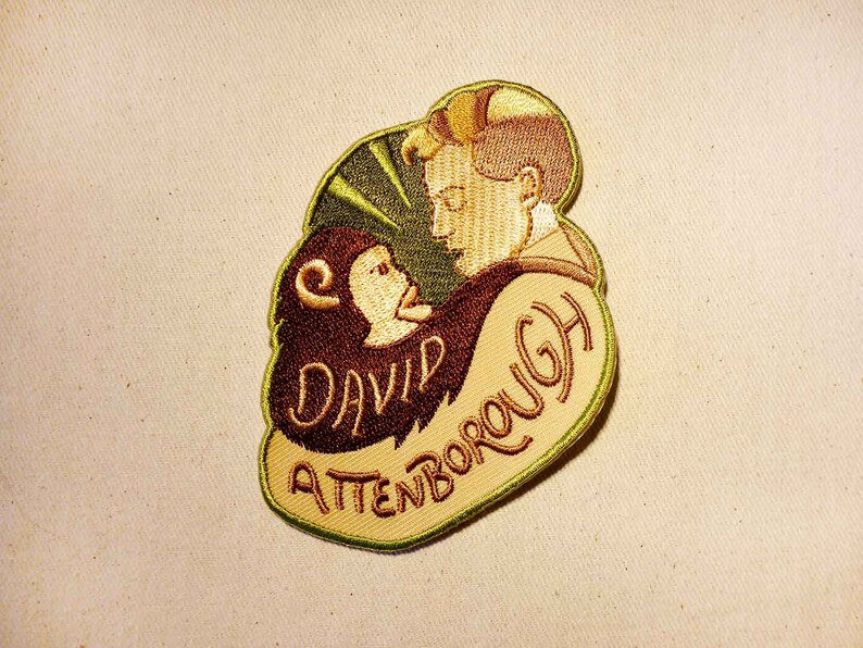 K&ouml;nnte beinhalten: Ein gestickter Aufn&auml;her in Gr&uuml;n, Braun und Beige mit einer Cartoon-Abbildung eines Mannes mit einem Affen auf der Schulter. Der Text "David Attenborough" ist in einer kursiv geschriebenen Schrift auf dem Aufn&auml;her gestickt.