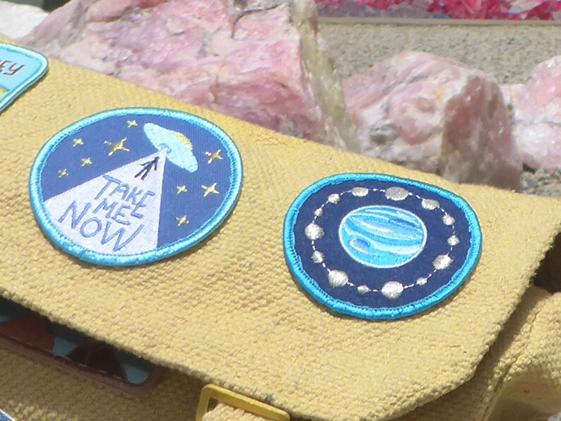 May include: Two embroidered patches on a beige fabric background. The left patch is blue with white text that says "Take Me Now" and a white alien spaceship with a beam of light pointing down. The right patch is blue with a silver circle around a blue planet.