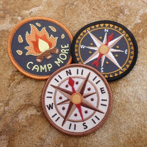 May include: Three embroidered patches, two with compass designs and one with a campfire and the text "Camp More".