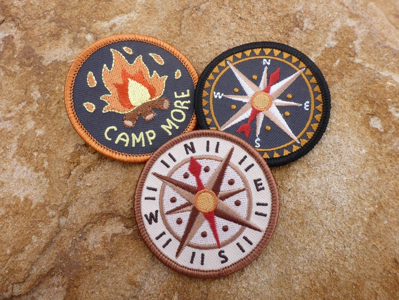 May include: Three embroidered patches, two with compass designs and one with a campfire and the text "Camp More".