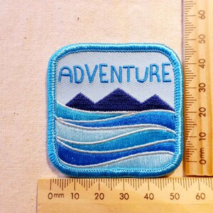 May include: Blue and white embroidered patch with the word "ADVENTURE" in white text. The patch features a blue mountain range with a blue and white wavy water design below.