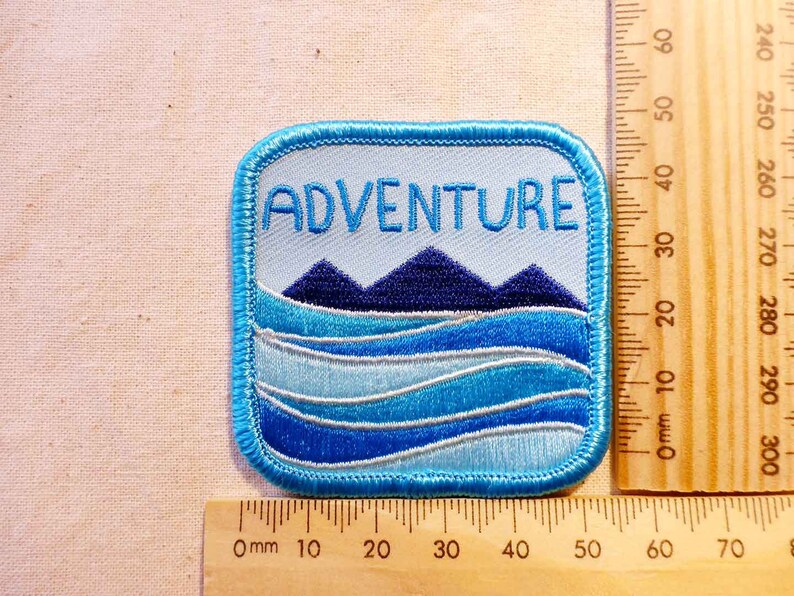 May include: Blue and white embroidered patch with the word "ADVENTURE" in white text. The patch features a blue mountain range with a blue and white wavy water design below.