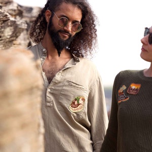 K&ouml;nnte beinhalten: Ein Mann und eine Frau stehen im Freien. Der Mann tr&auml;gt ein hellbraunes Hemd mit einem Aufn&auml;her auf der linken Brustseite. Die Frau tr&auml;gt einen dunkelgr&uuml;nen Pullover mit mehreren Aufn&auml;hern auf der Vorderseite.