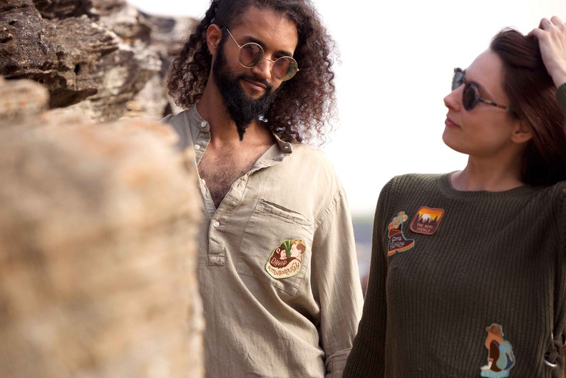 K&ouml;nnte beinhalten: Ein Mann und eine Frau stehen im Freien. Der Mann tr&auml;gt ein hellbraunes Hemd mit einem Aufn&auml;her auf der linken Brustseite. Die Frau tr&auml;gt einen dunkelgr&uuml;nen Pullover mit mehreren Aufn&auml;hern auf der Vorderseite.