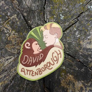 K&ouml;nnte beinhalten: Gestickter Aufn&auml;her mit einer Cartoon-Illustration von David Attenborough mit einem Affen, mit dem Text "David Attenborough" in braunem Garn.