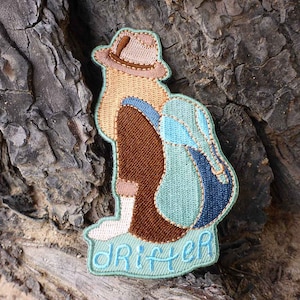 May include: Embroidered patch of a person wearing a brown hat and a blue backpack, with the word "Drifter" stitched below.