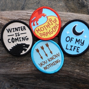 Puede incluir: Cuatro parches bordados para planchar con diferentes diseños y textos. Los parches están sobre una superficie de madera. Los parches son azules, amarillos, rojos y blancos. El texto en los parches dice "Winter is Coming", "Mother of Dragons", "Of My Life", y "You Know Nothing".