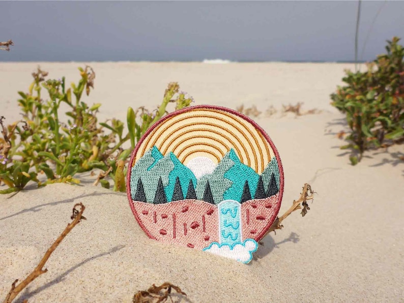 K&ouml;nnte beinhalten: Ein runder gestickter Aufn&auml;her mit einer Bergszene. Der Aufn&auml;her zeigt einen Sonnenuntergang mit einem Regenbogen, Bergen, B&auml;umen und einem Wasserfall. Der Aufn&auml;her befindet sich auf einem Sandstrand.