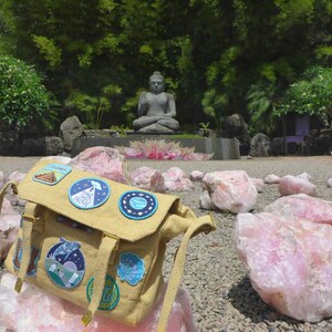May include: A tan canvas messenger bag with a variety of colourful patches, including a UFO, a planet, and a pyramid. The bag is sitting on a pile of pink quartz crystals in a garden setting with a statue of Buddha in the background.