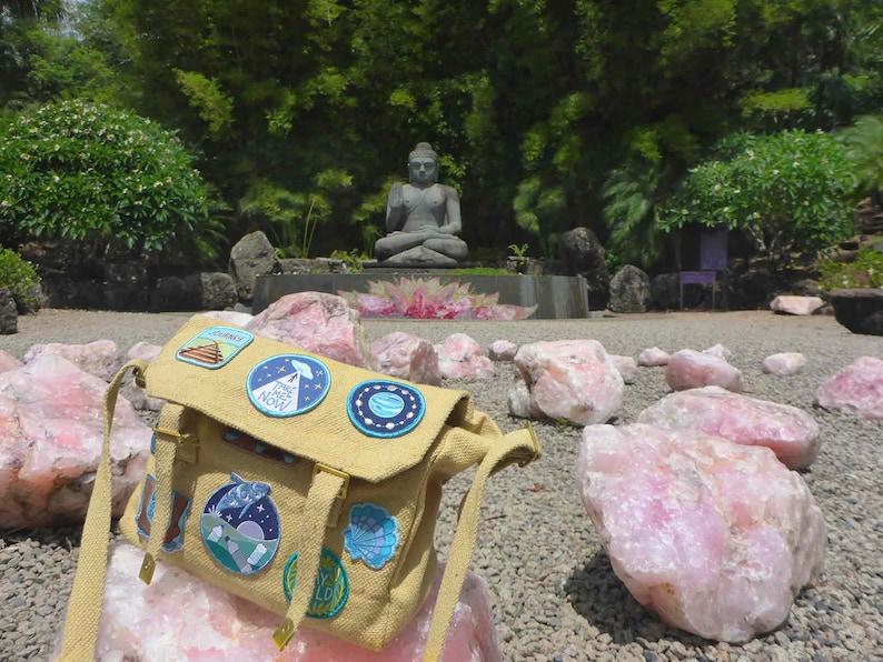 May include: A tan canvas messenger bag with a variety of colourful patches, including a UFO, a planet, and a pyramid. The bag is sitting on a pile of pink quartz crystals in a garden setting with a statue of Buddha in the background.