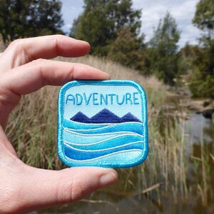 May include: A blue and white embroidered patch with the word "ADVENTURE" in white text. The patch features a blue mountain range and blue waves.