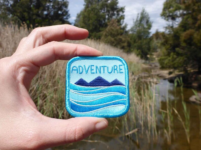 May include: A blue and white embroidered patch with the word "ADVENTURE" in white text. The patch features a blue mountain range and blue waves.