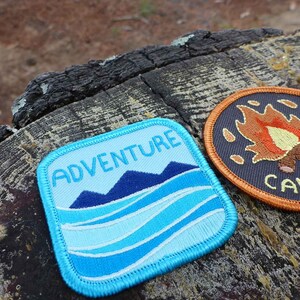 May include: Two embroidered patches, one blue with the text "ADVENTURE" and a mountain and wave design, and the other orange with the text "CAMP MORE" and a campfire design.