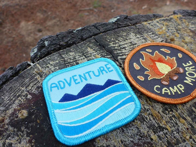 May include: Two embroidered patches, one blue with the text "ADVENTURE" and a mountain and wave design, and the other orange with the text "CAMP MORE" and a campfire design.