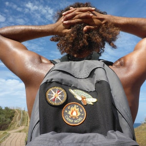 May include: A person with curly hair wearing a gray backpack with three patches. The patches are a compass, a butterfly, and a campfire with the text "Camp More".
