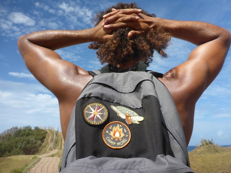 May include: A person with curly hair wearing a gray backpack with three patches. The patches are a compass, a butterfly, and a campfire with the text "Camp More".