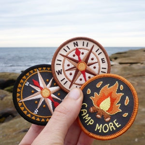 May include: Three embroidered patches with a camping theme. The patches are round and feature a compass, a campfire, and the text "Camp More".