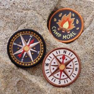 May include: Three embroidered patches with a camping theme. The first patch is black with a compass rose in the center. The second patch is brown with a compass rose in the center. The third patch is black with a campfire and the text "Camp More".