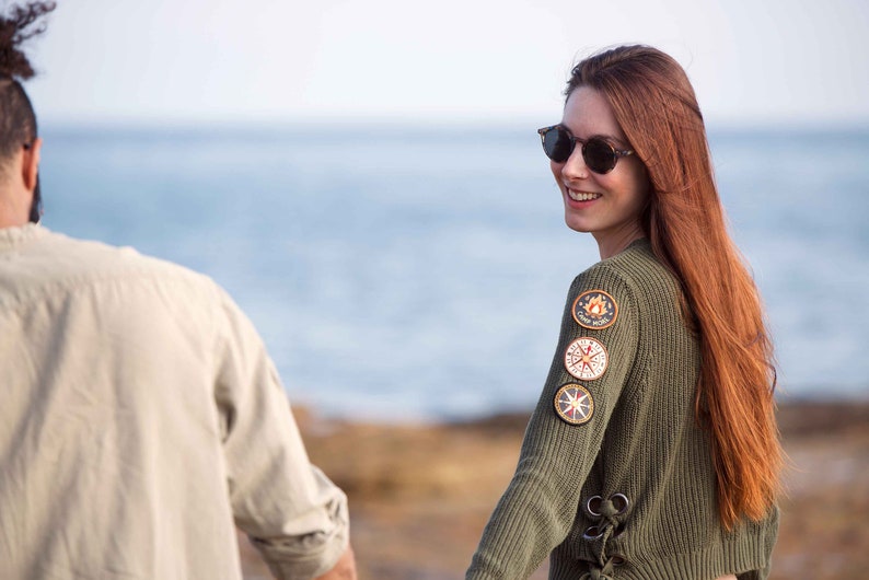May include: A woman with long red hair wearing a green sweater with patches on the sleeve. She is smiling and looking over her shoulder. The background is a blurry image of the ocean.
