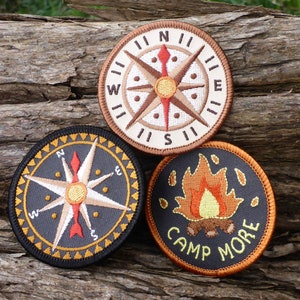May include: Three embroidered patches with a camping theme. The first patch is a compass with a brown background and a white compass rose. The second patch is a compass with a black background and a white compass rose. The third patch is a campfire with a black background and orange flames. The text on the third patch reads "Camp More".