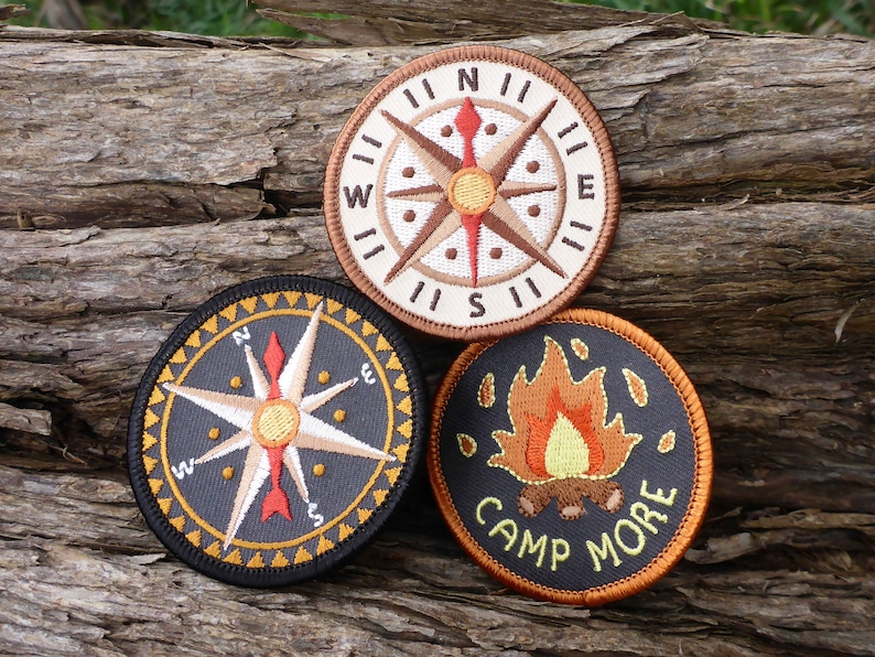 May include: Three embroidered patches with a camping theme. The first patch is a compass with a brown background and a white compass rose. The second patch is a compass with a black background and a white compass rose. The third patch is a campfire with a black background and orange flames. The text on the third patch reads "Camp More".