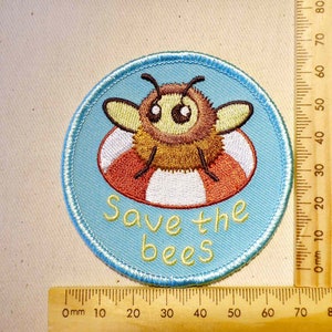 May include: A blue and white embroidered patch with a cartoon bee wearing a red and white life preserver. The text "Save the bees" is written below the bee.