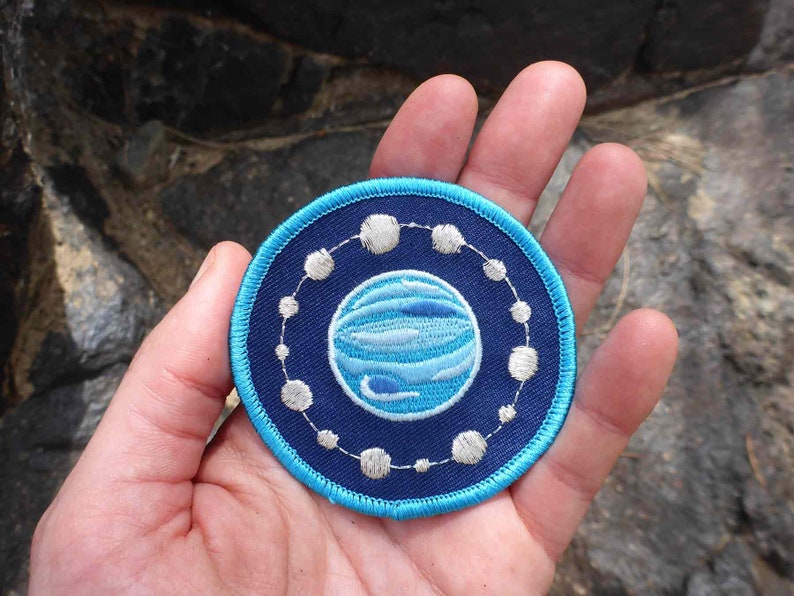 May include: A blue and white embroidered patch featuring a planet design with a ring of white stars around it.