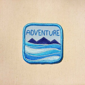 May include: Blue and white embroidered patch with the word "ADVENTURE" in white text and a blue mountain range and waves design.