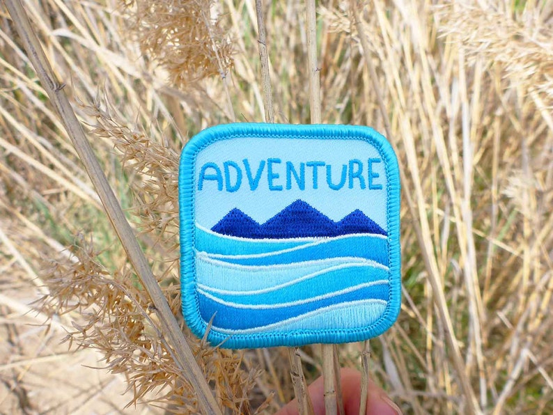 May include: A blue and white embroidered patch with the word "ADVENTURE" in white text and a blue mountain range and blue waves design.