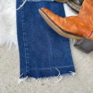 K&ouml;nnte beinhalten: Eine blaue Jeans mit ausgefransten S&auml;umen und einem Used-Look. Die Jeans liegt auf einer wei&szlig;en, flauschigen Oberfl&auml;che mit einem Paar braunen Leder-Cowboots.