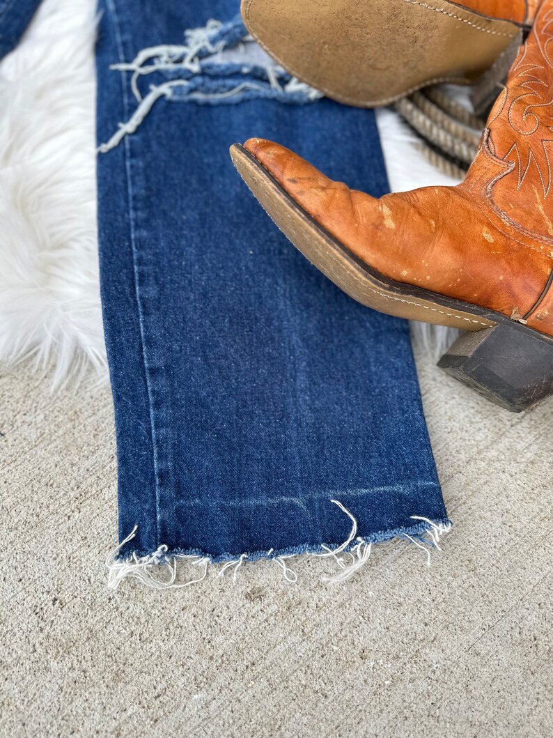 K&ouml;nnte beinhalten: Eine blaue Jeans mit ausgefransten S&auml;umen und einem Used-Look. Die Jeans liegt auf einer wei&szlig;en, flauschigen Oberfl&auml;che mit einem Paar braunen Leder-Cowboots.